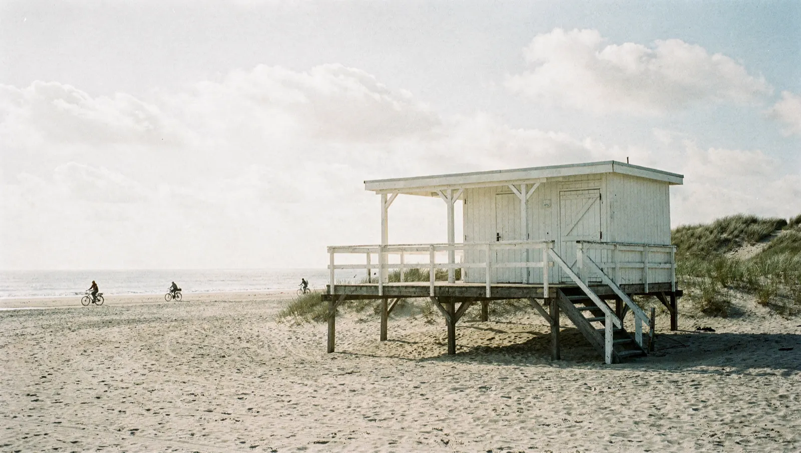 A white beach cabin on stilts in the dunes, with cyclists along the waterline behind it