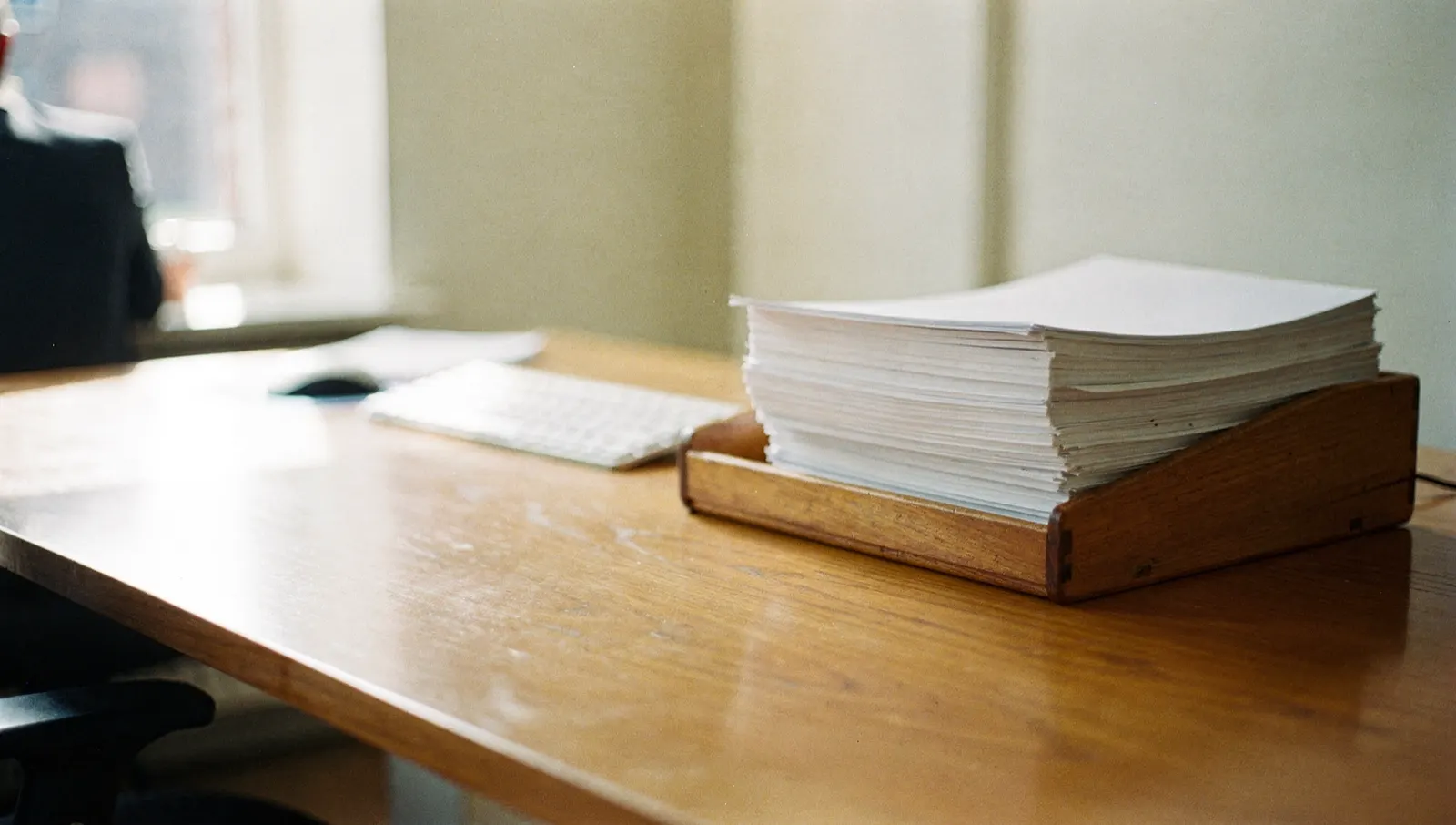 A wooden paper tray heaped with plain sheets beside a keyboard in morning light