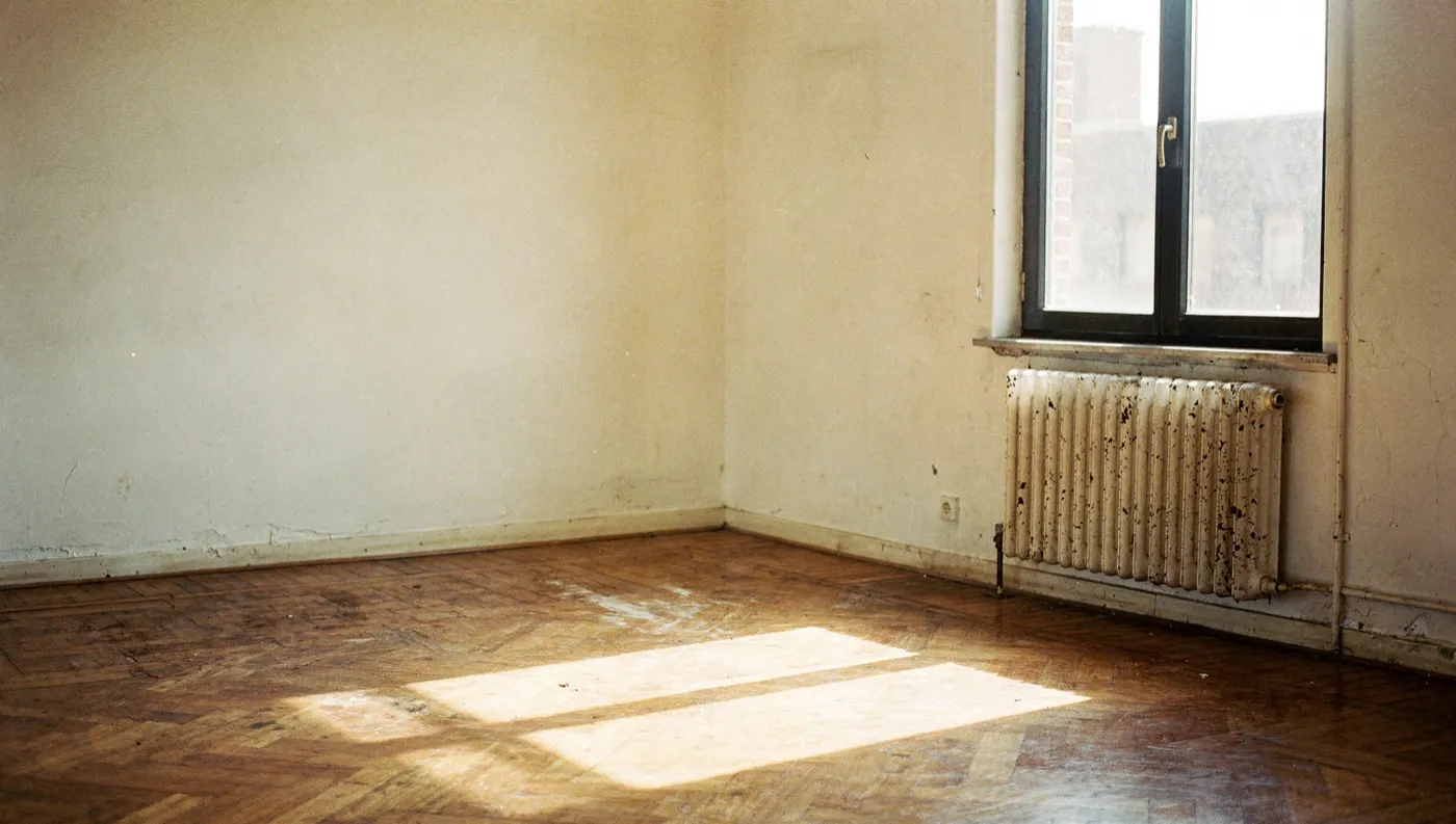 An empty, sunlit but dated Flemish room with a worn parquet floor and an old radiator