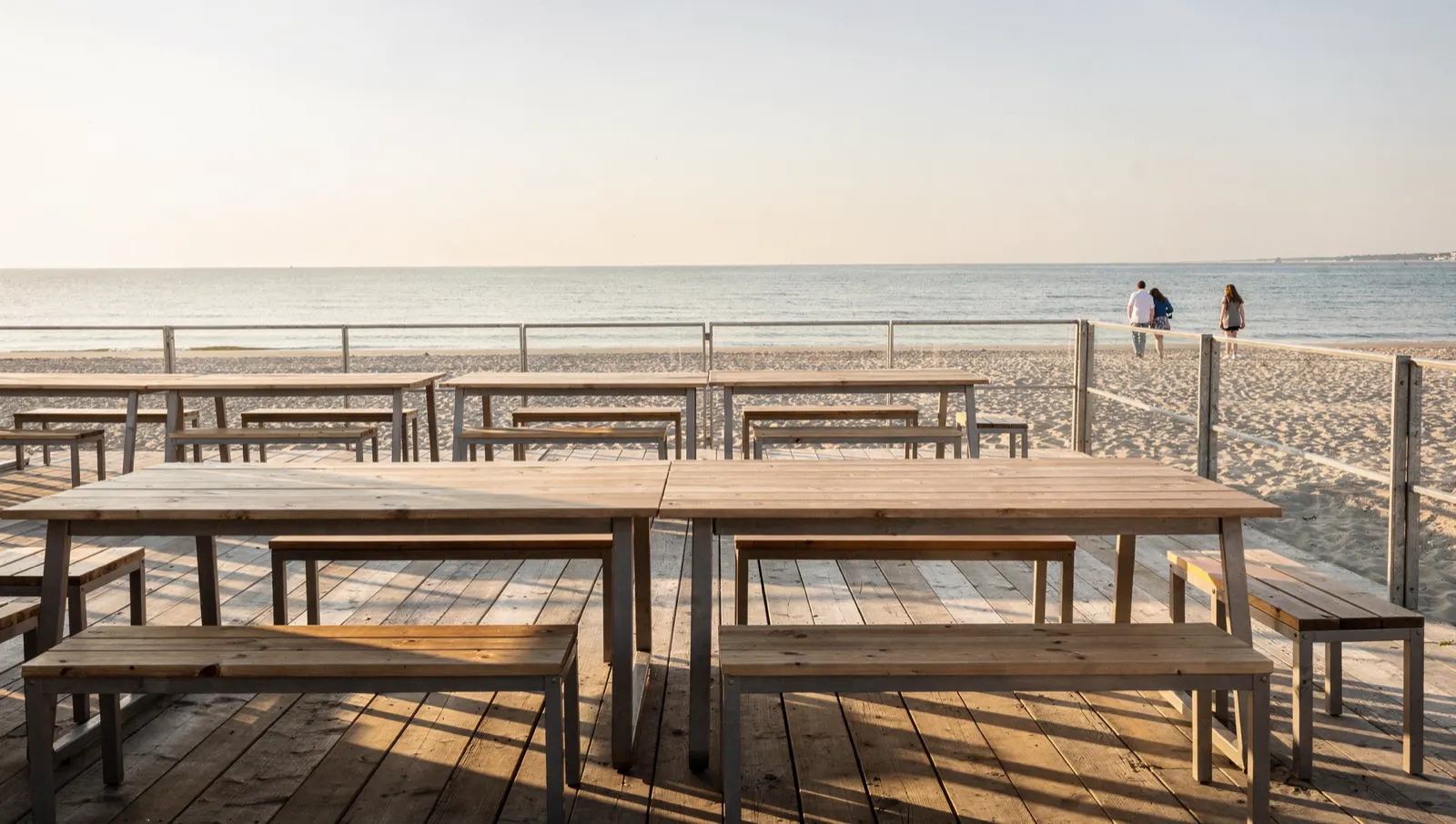 A beach bar terrace with long timber tables in low afternoon sun, the sea beyond