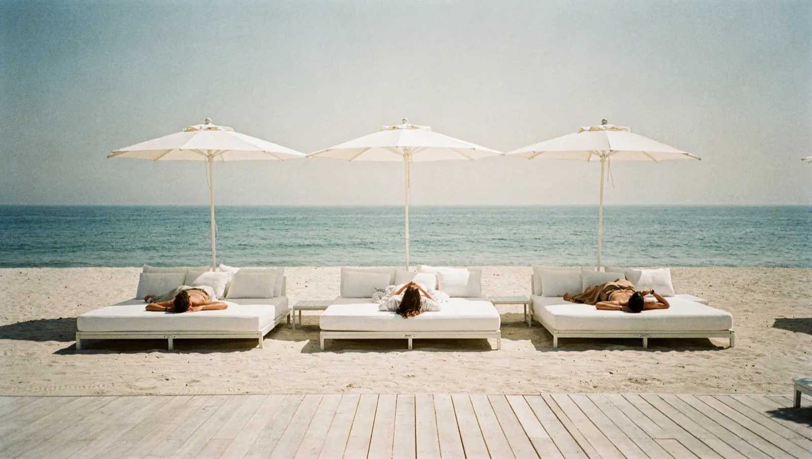 The front row of a beach club, wide white daybeds under parasols at the edge of the sand