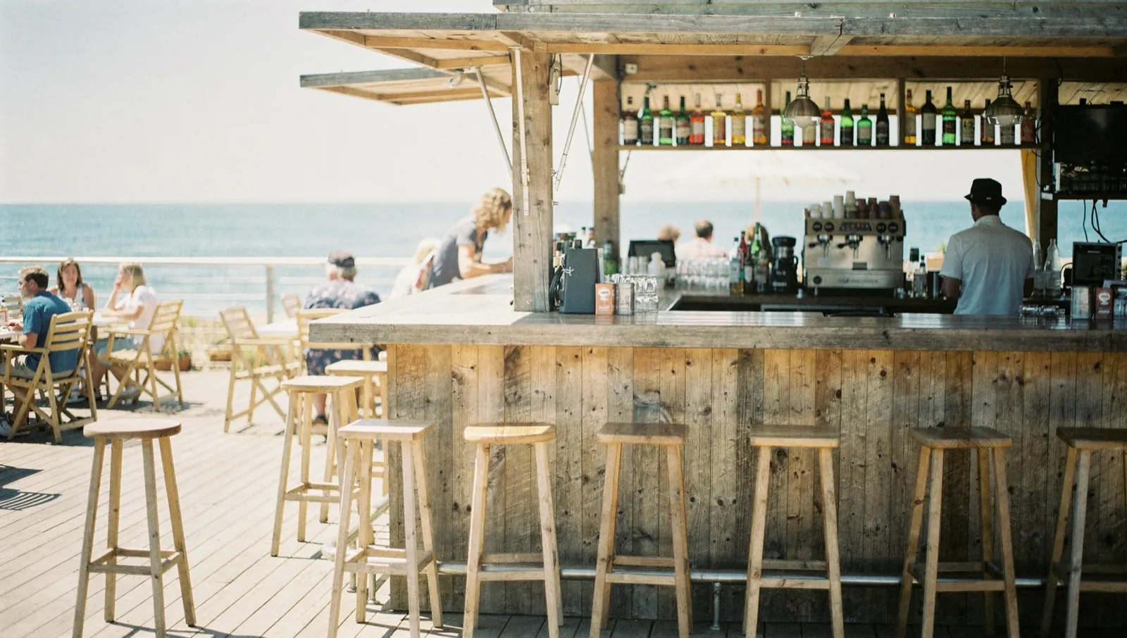 The counter of a beach bar on a busy afternoon, a full terrace and the sea beyond the open frontage