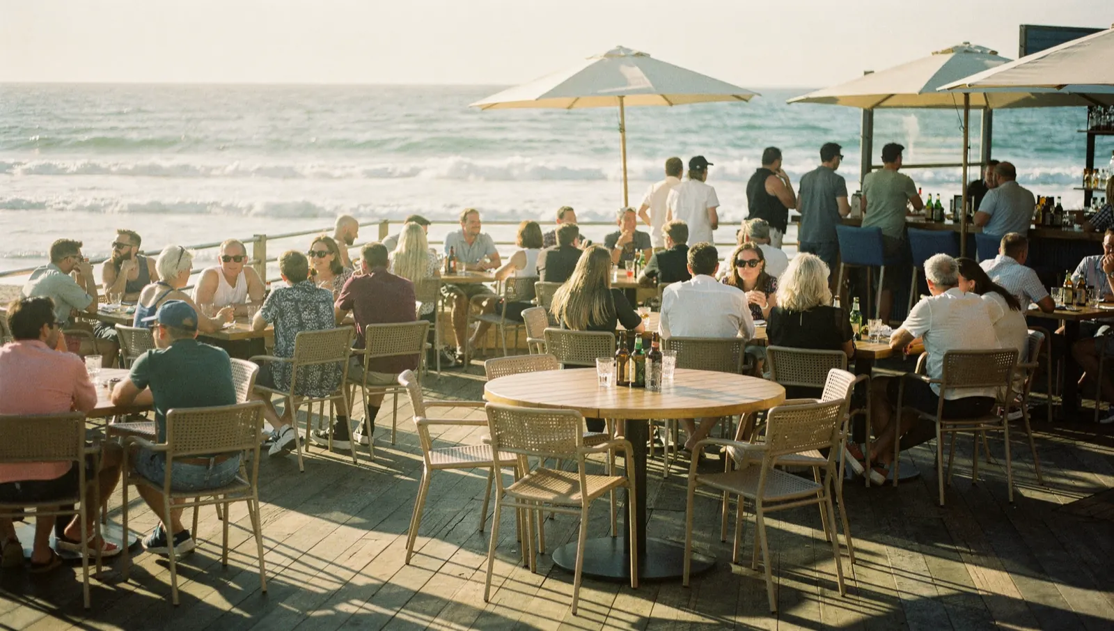 A beach bar terrace at its busiest, every table taken and people standing along the seaward edge