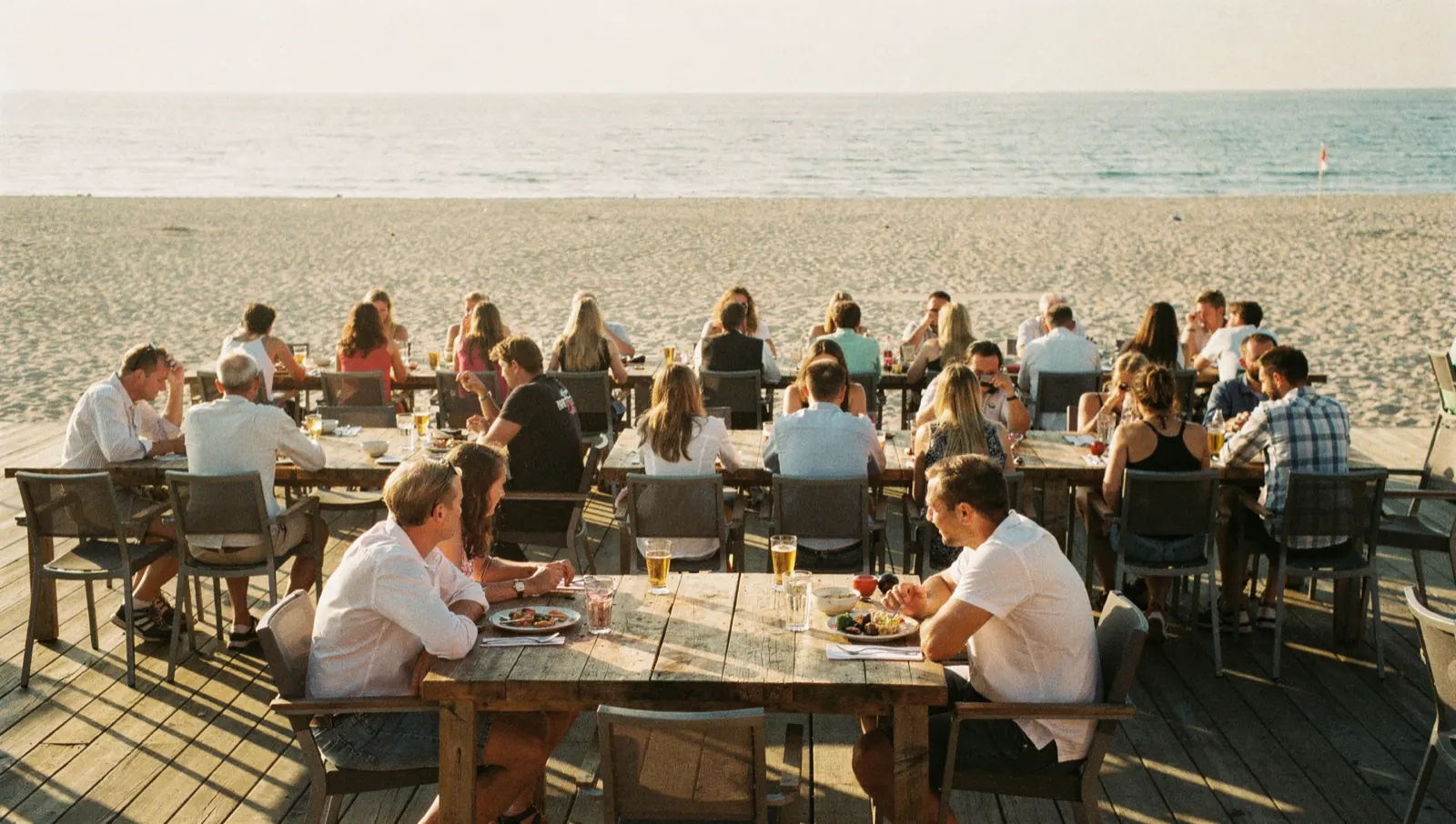 A full beach bar terrace at golden hour, guests seated along long timber tables with the sea behind